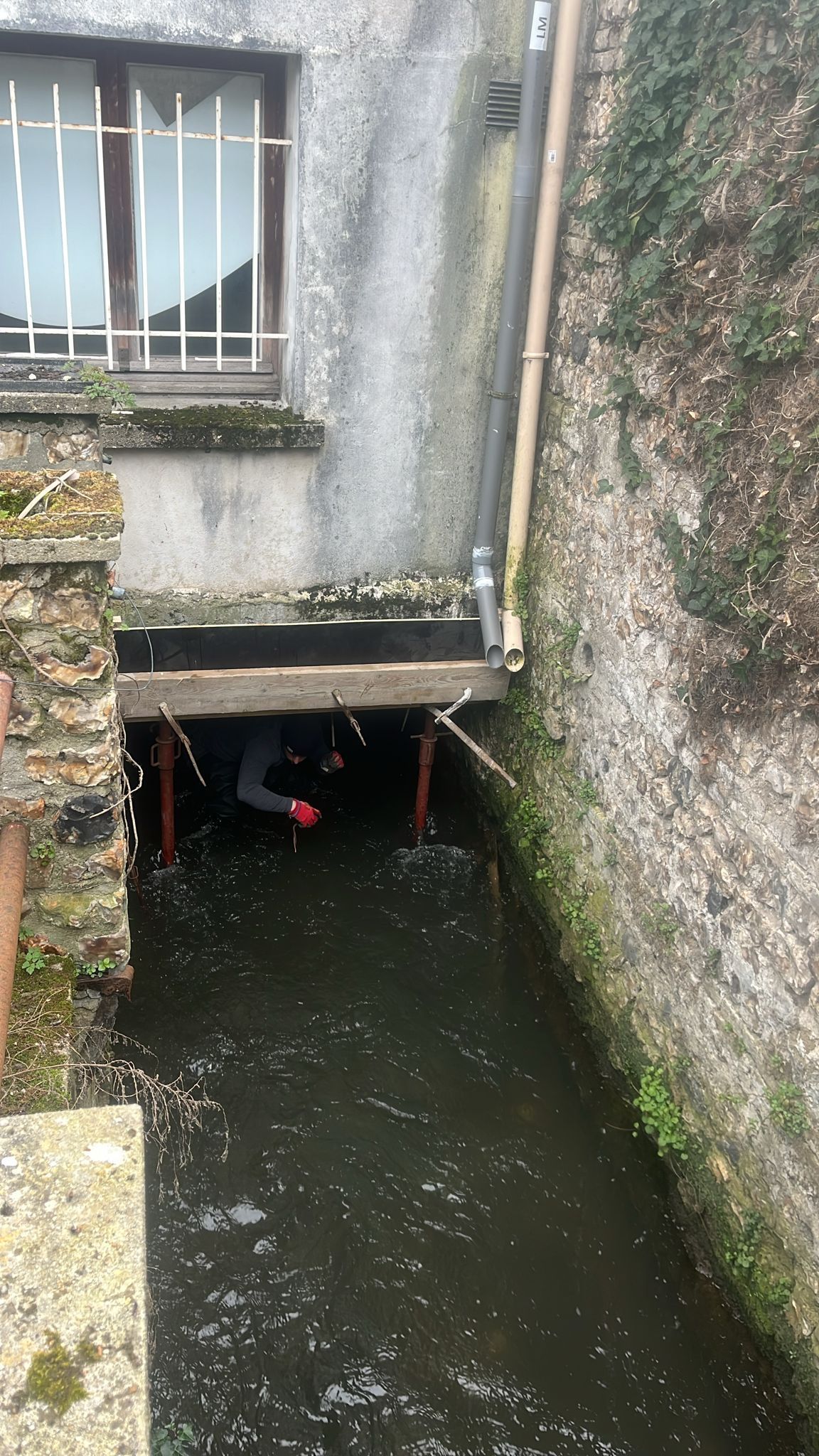 Ruelle étroite et inondée, longeant de vieux murs de pierre et une fenêtre, où l'eau sombre stagne sous un rebord bas.