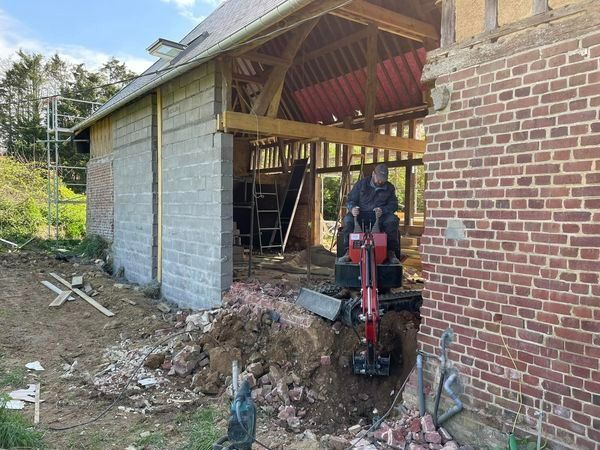 Maison en briques partiellement construite, avec un mur ouvert, des débris de construction et un ouvrier utilisant un marteau-piqueur rouge.