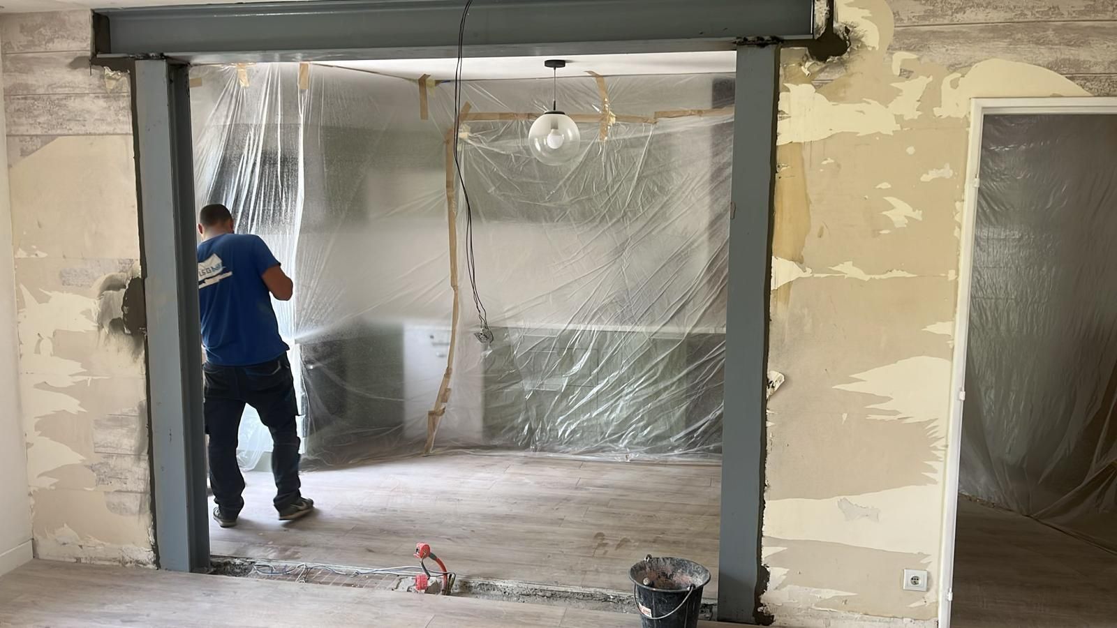 Un ouvrier en chemise bleue manipule un cadre de porte dans une pièce en rénovation.