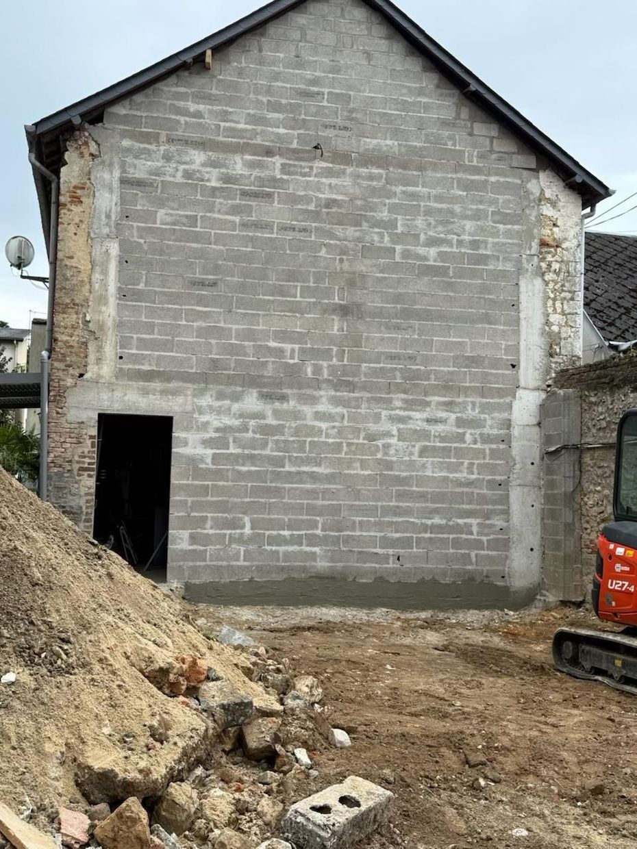 Bâtiment en béton gris inachevé, avec une grande porte, des tas de terre devant et une petite excavatrice sur la droite.