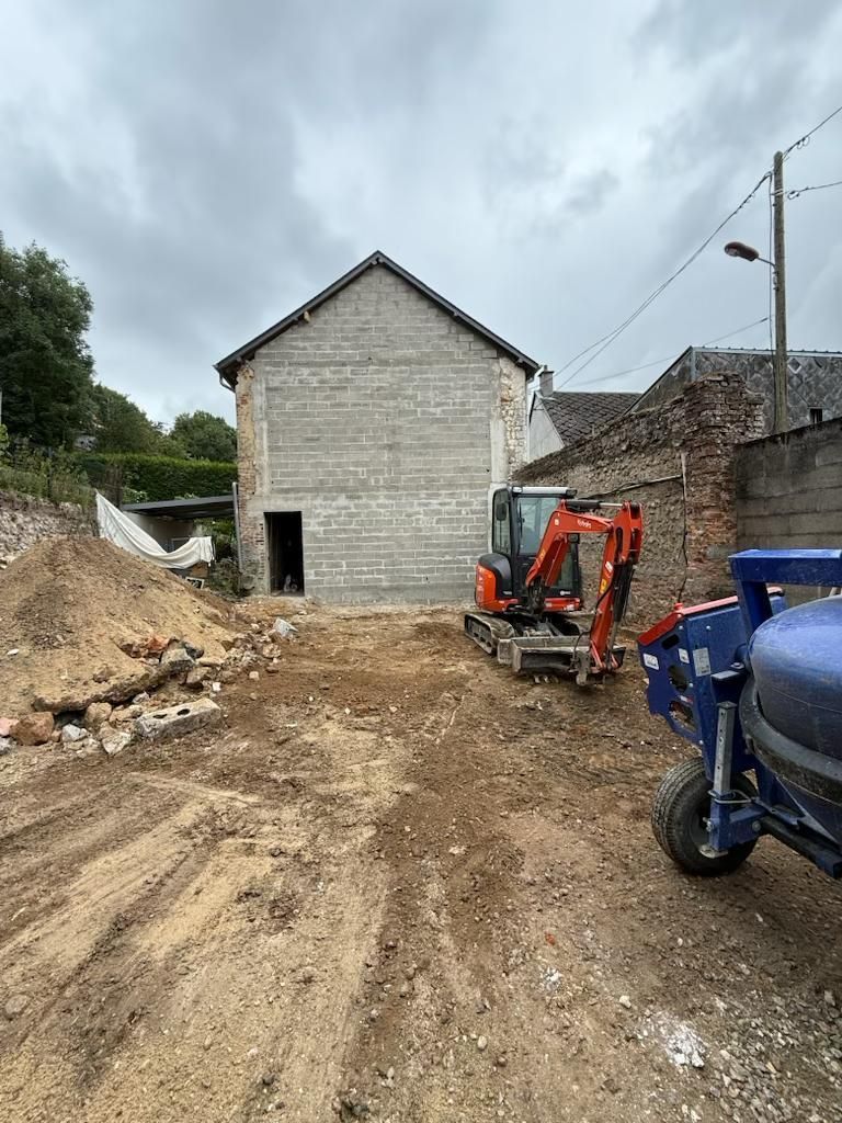 Chantier de construction avec un bâtiment en pierre, une excavatrice et un véhicule utilitaire bleu sur un chemin de terre boueux sous un ciel nuageux.