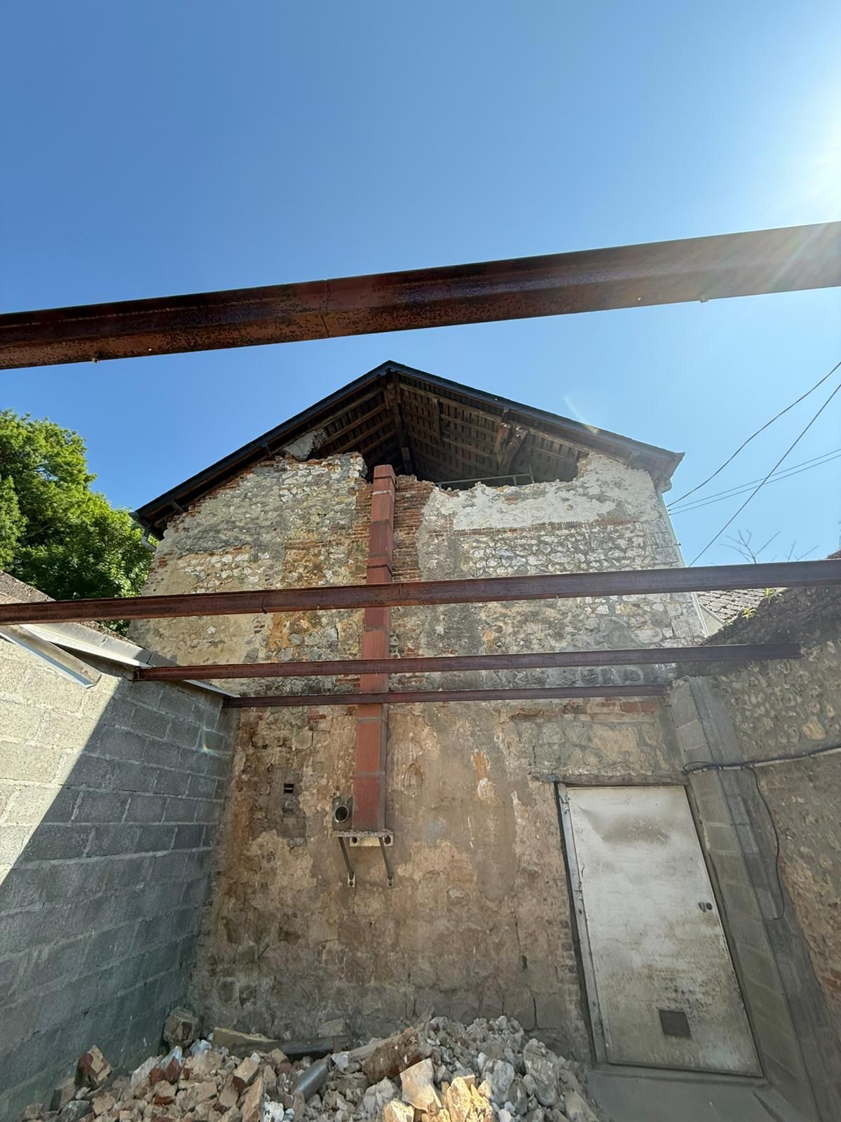 Vieux bâtiment en pierre avec une porte en métal, se détachant sur un ciel bleu avec des poutres rouillées au premier plan.