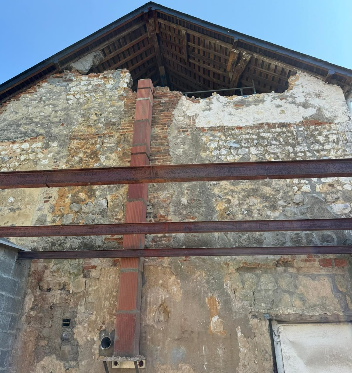 Vieux mur de pierre avec cheminée en briques rouges et toit apparent sous un ciel bleu