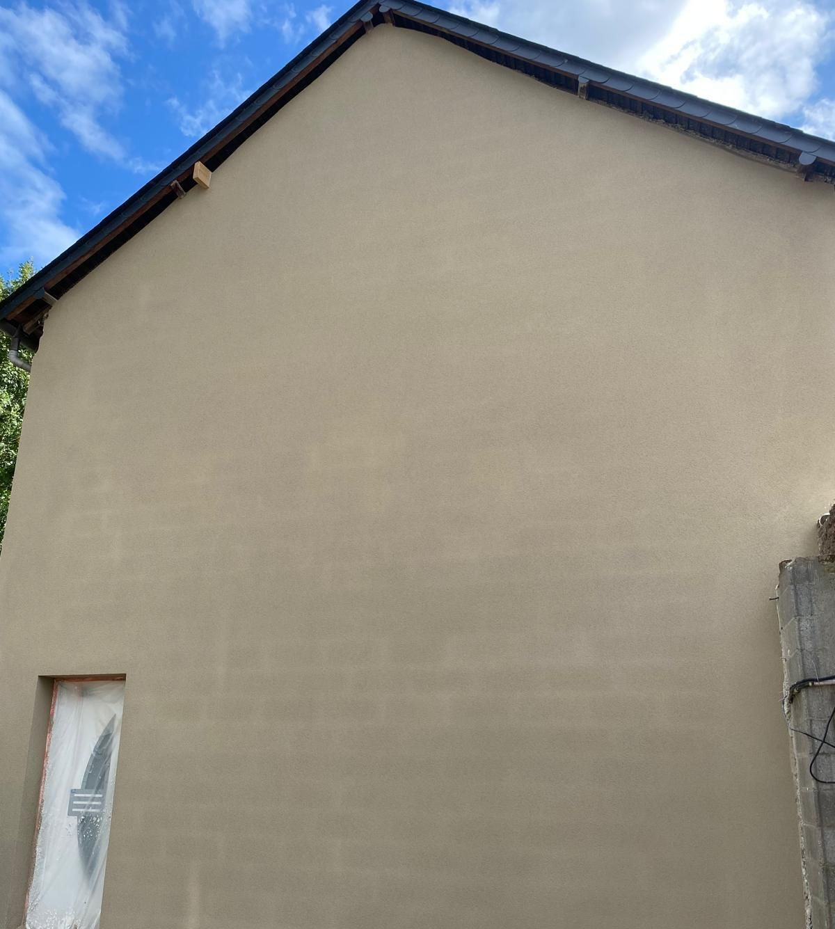 Mur d'une maison en stuc beige et toit à pignon se détachant sur un ciel bleu partiellement nuageux
