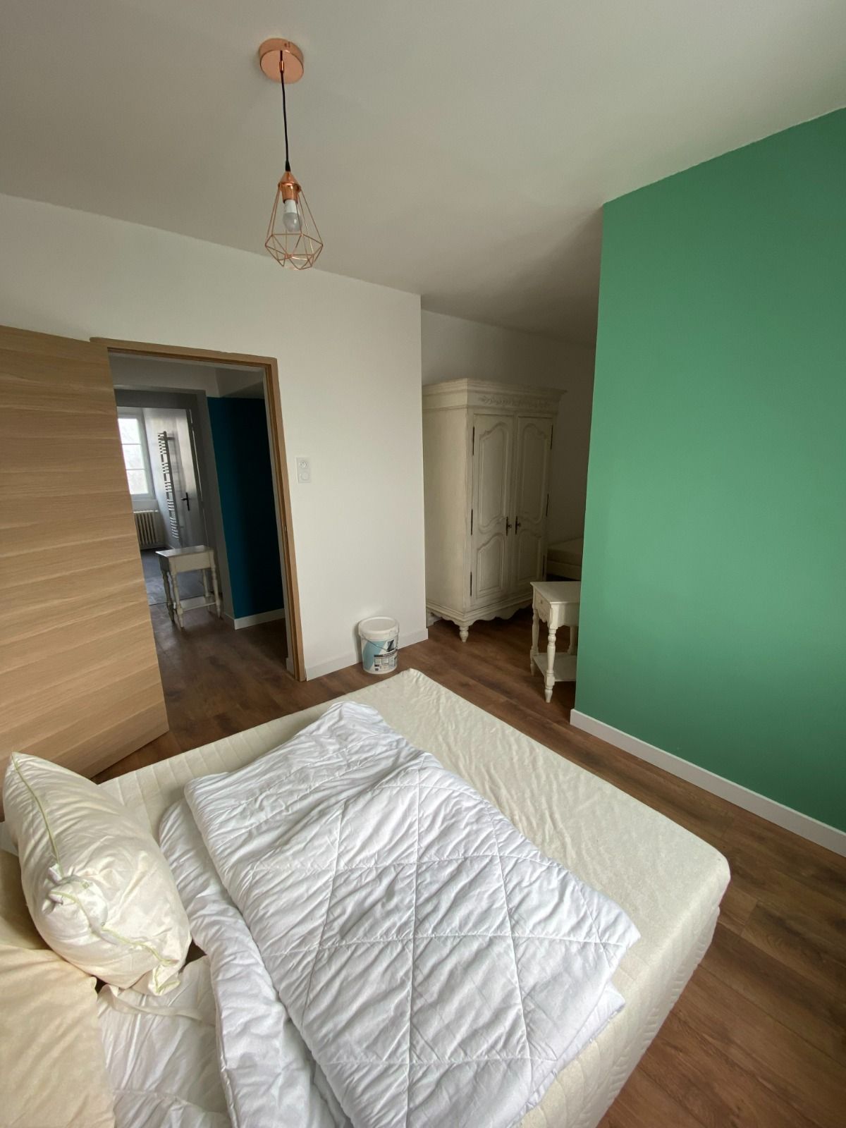 Chambre lumineuse avec un lit blanc, un mur d'accent vert, un parquet et une porte ouverte sur un couloir