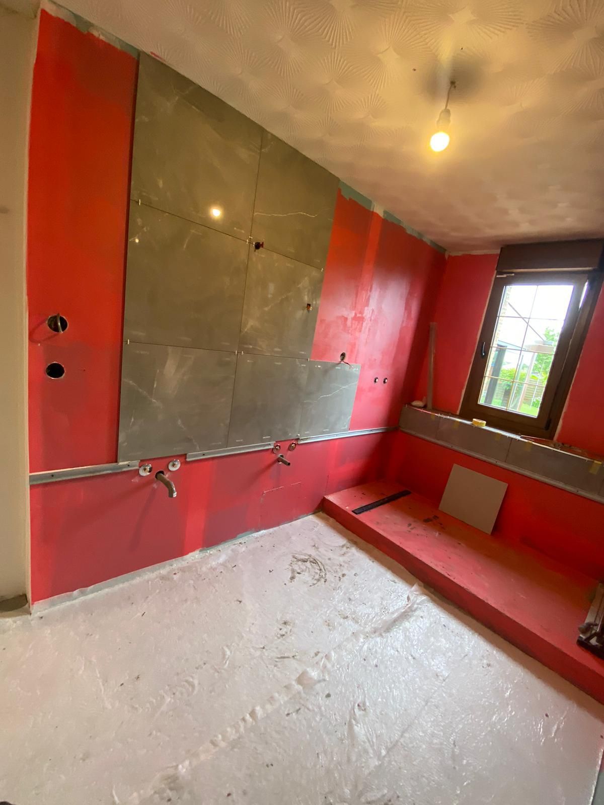 Petite pièce rouge et grise inachevée avec carrelage mural, fenêtre et banquette.