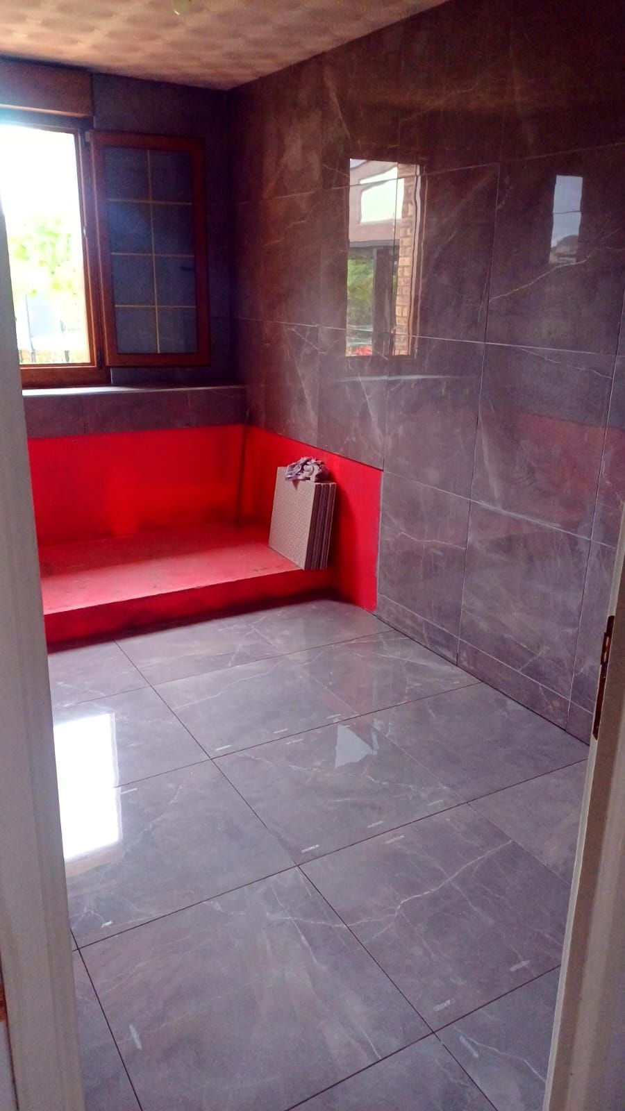 Petite pièce carrelée avec une banquette rouge intégrée, une fenêtre et des murs gris brillants.