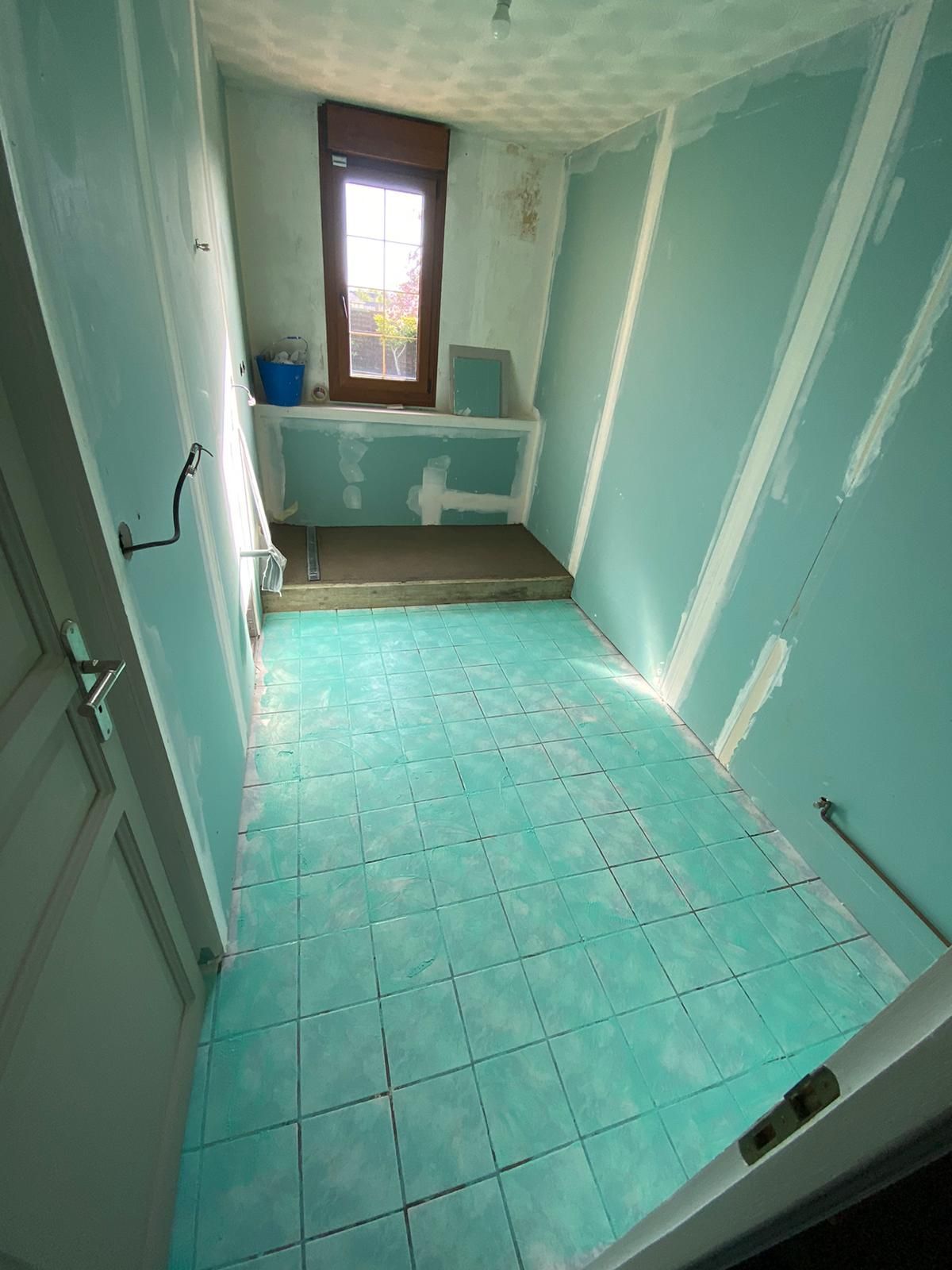 Salle de bain étroite carrelée de turquoise, avec des cloisons sèches non finies et une fenêtre au fond.