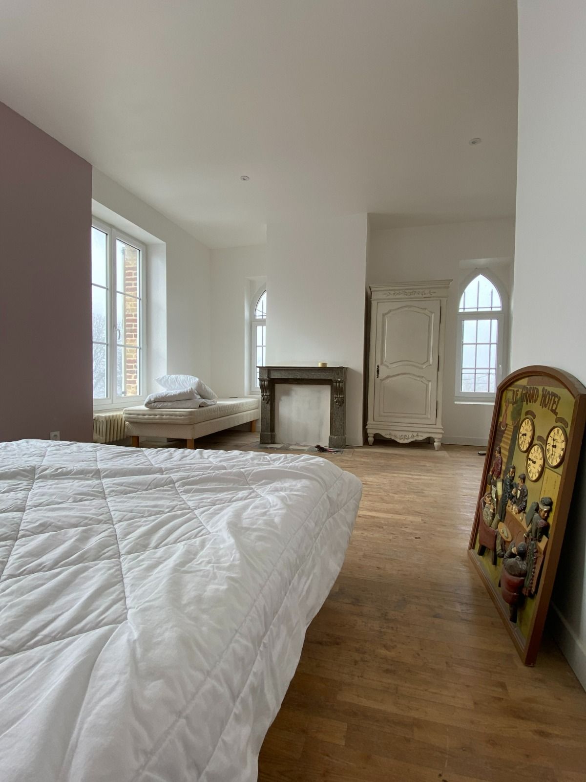 Chambre lumineuse avec lit blanc, parquet, cheminée, hautes fenêtres et un tableau coloré encadré, légèrement incliné.
