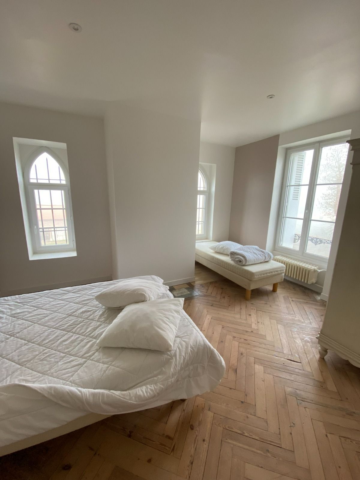 Chambre lumineuse avec deux lits défaits, murs blancs, parquet et grandes fenêtres laissant entrer la lumière naturelle.