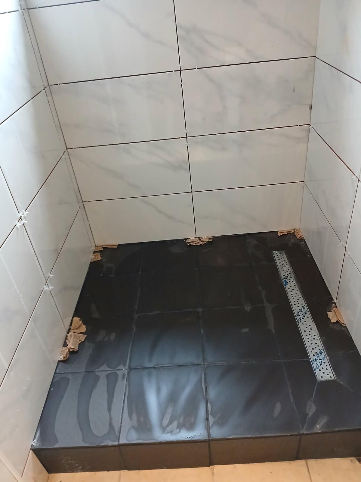 Petit coin de douche carrelé avec receveur de douche et bonde noirs, présentant des traces d'eau et des débris.
