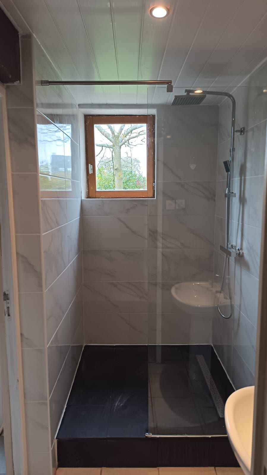 Salle de bain moderne avec douche à paroi de verre, murs carrelés blancs, sol foncé et petite fenêtre.