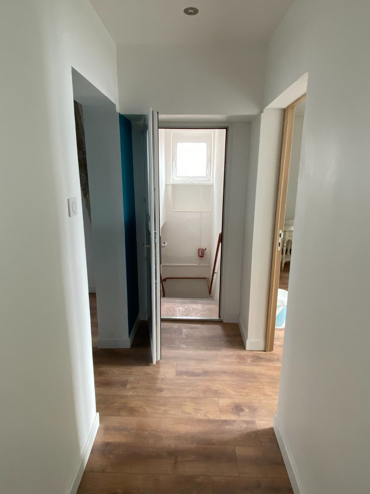 Couloir étroit aux murs blancs, au sol en bois et aux portes menant à une petite pièce avec une fenêtre.