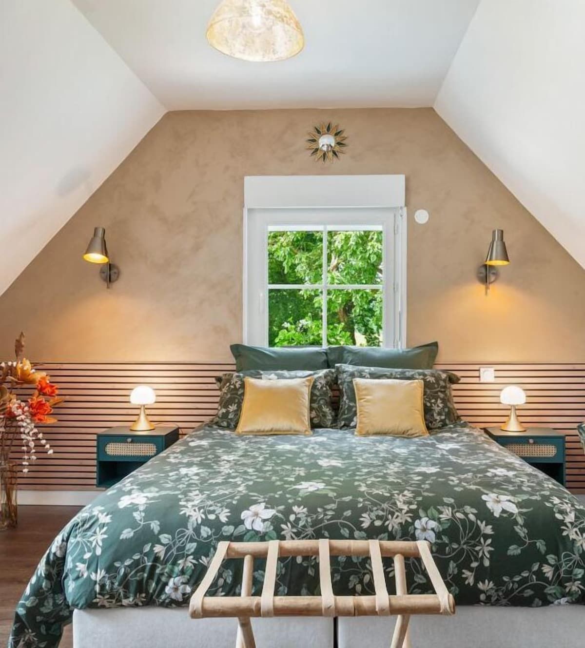 Chambre mansardée confortable avec linge de lit à motifs floraux verts, murs aux tons chauds et une fenêtre centrée au-dessus du lit.