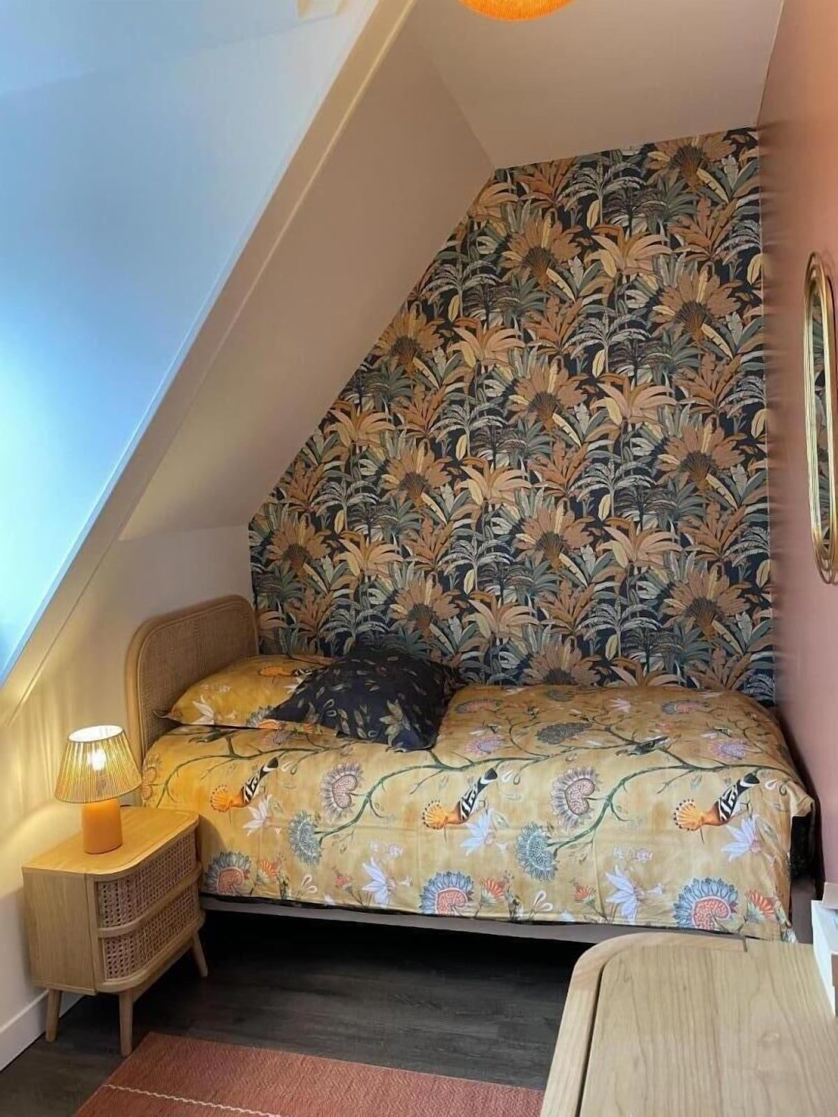Chambre mansardée confortable avec couvre-lit à motifs, tête de lit fleurie et lampe de chevet sous un plafond mansardé