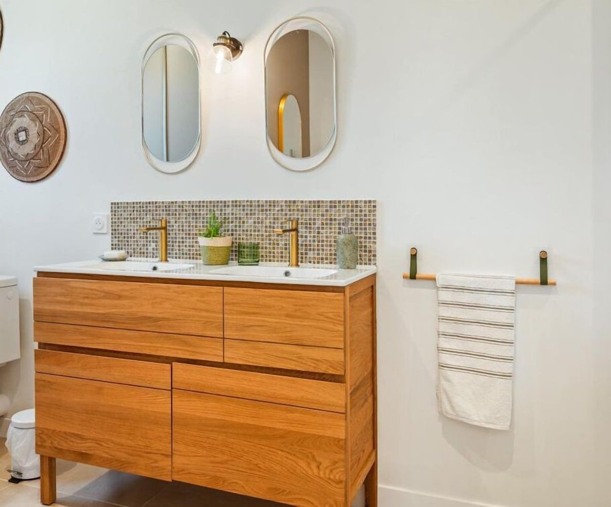 Meuble-lavabo moderne avec armoire en bois, miroirs ronds, crédence carrelée et serviettes blanches accrochées au mur.