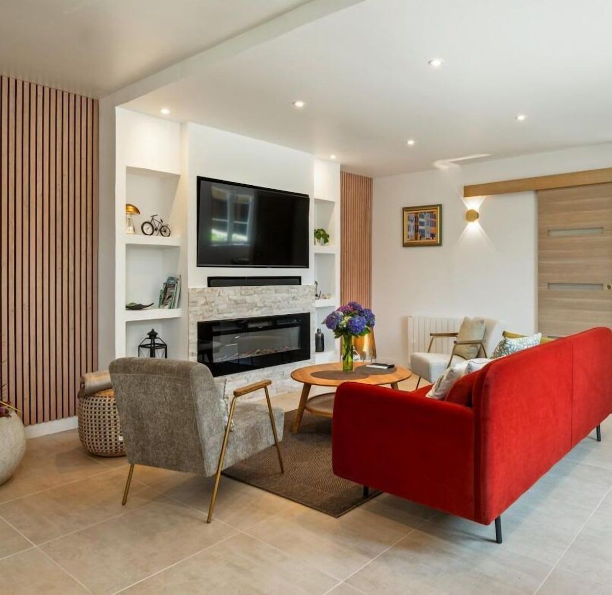 Salon moderne avec canapé rouge, fauteuil gris, télévision murale, cheminée et table basse fleurie