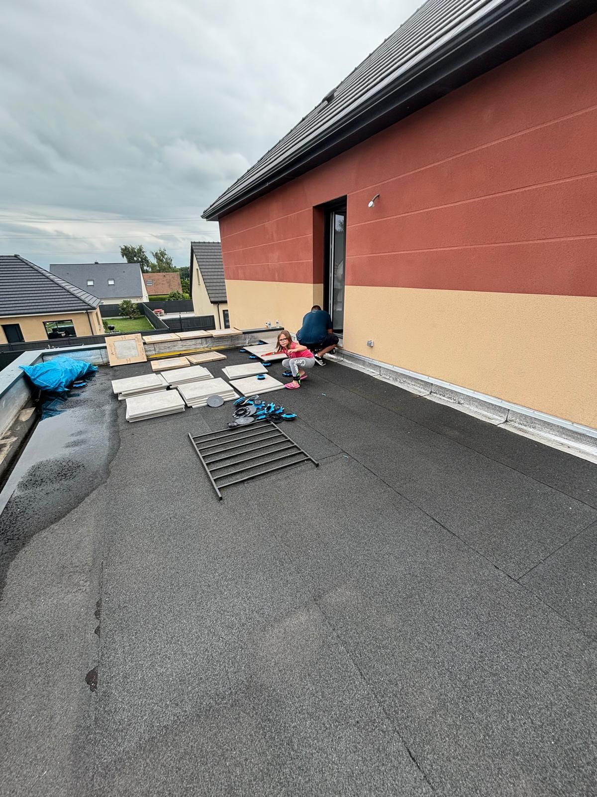 Un couvreur travaille sur un toit plat à côté d'un bâtiment rouge et crème, avec ses outils et matériaux disposés à proximité.