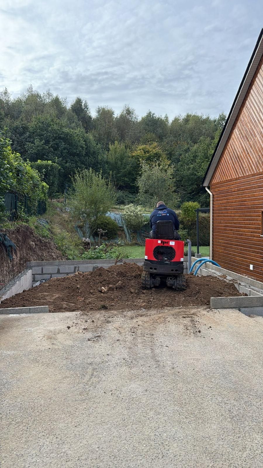 Une personne sur un quad rouge sur une allée de gravier à côté d'une maison en briques, avec une colline boisée en arrière-plan.