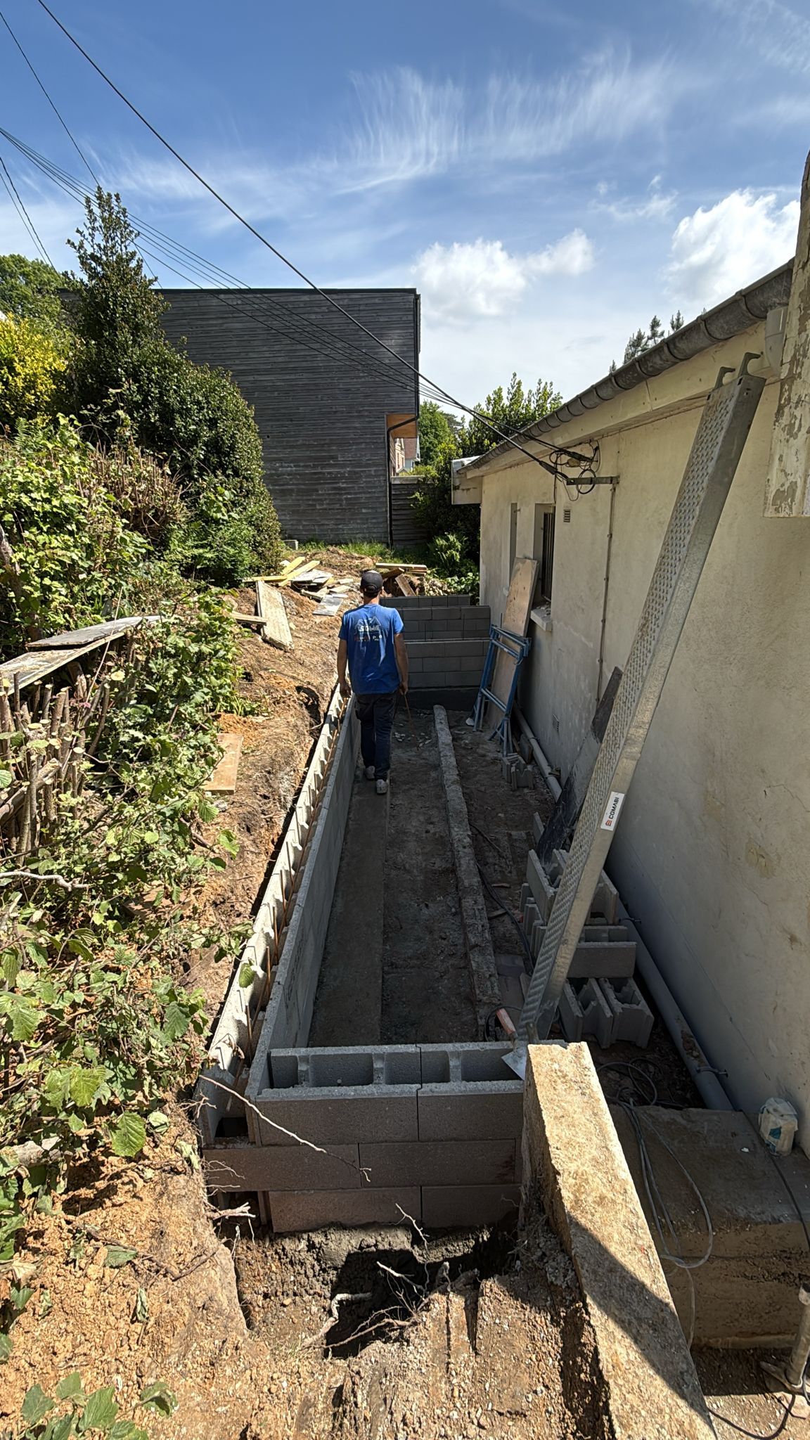 Tranchée étroite à l'extérieur, jouxtant une maison, avec des parpaings, du gravier et une personne en veste bleue travaillant à l'intérieur.