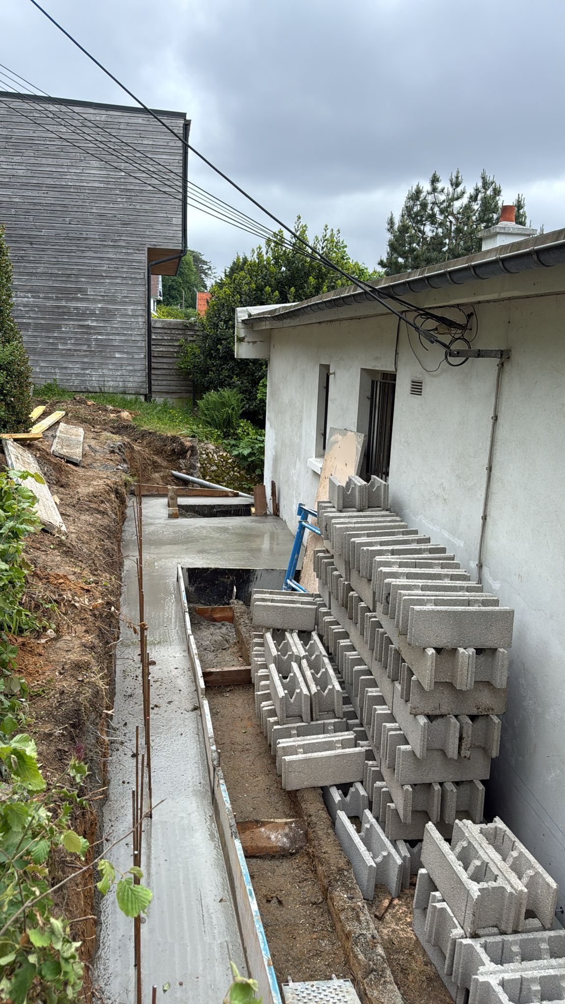 Étroite zone de construction extérieure jouxtant un bâtiment blanc, avec des parpaings empilés et une tranchée pour la gouttière.