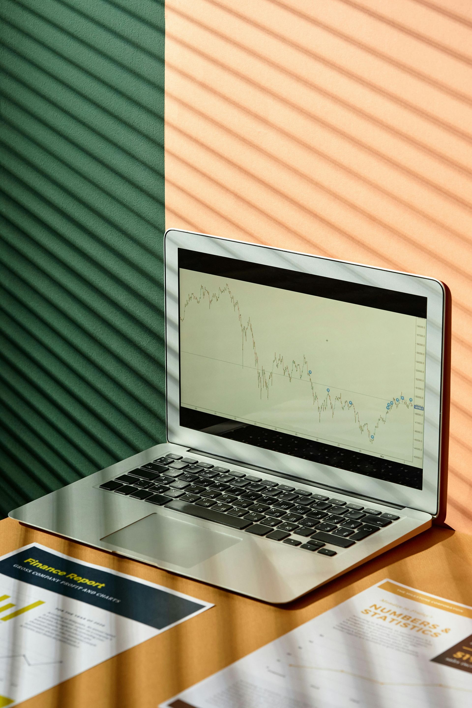 Laptop displaying a graph, on a desk with papers. Shadow from blinds on wall.