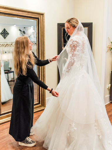 Woman in wedding dress, being assisted by another person in a boutique.