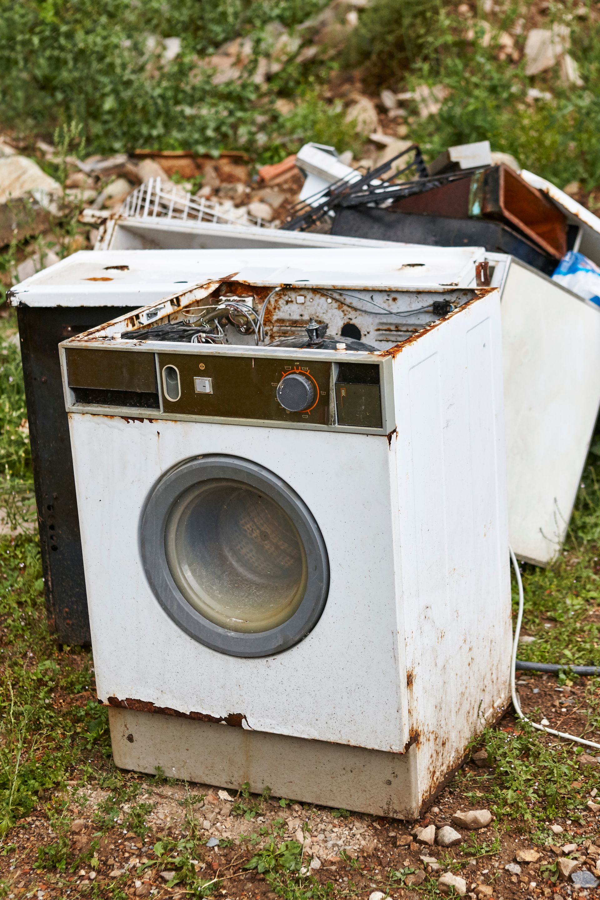 Broken white washing machine in a grassy outdoor area, with rust and debris around it