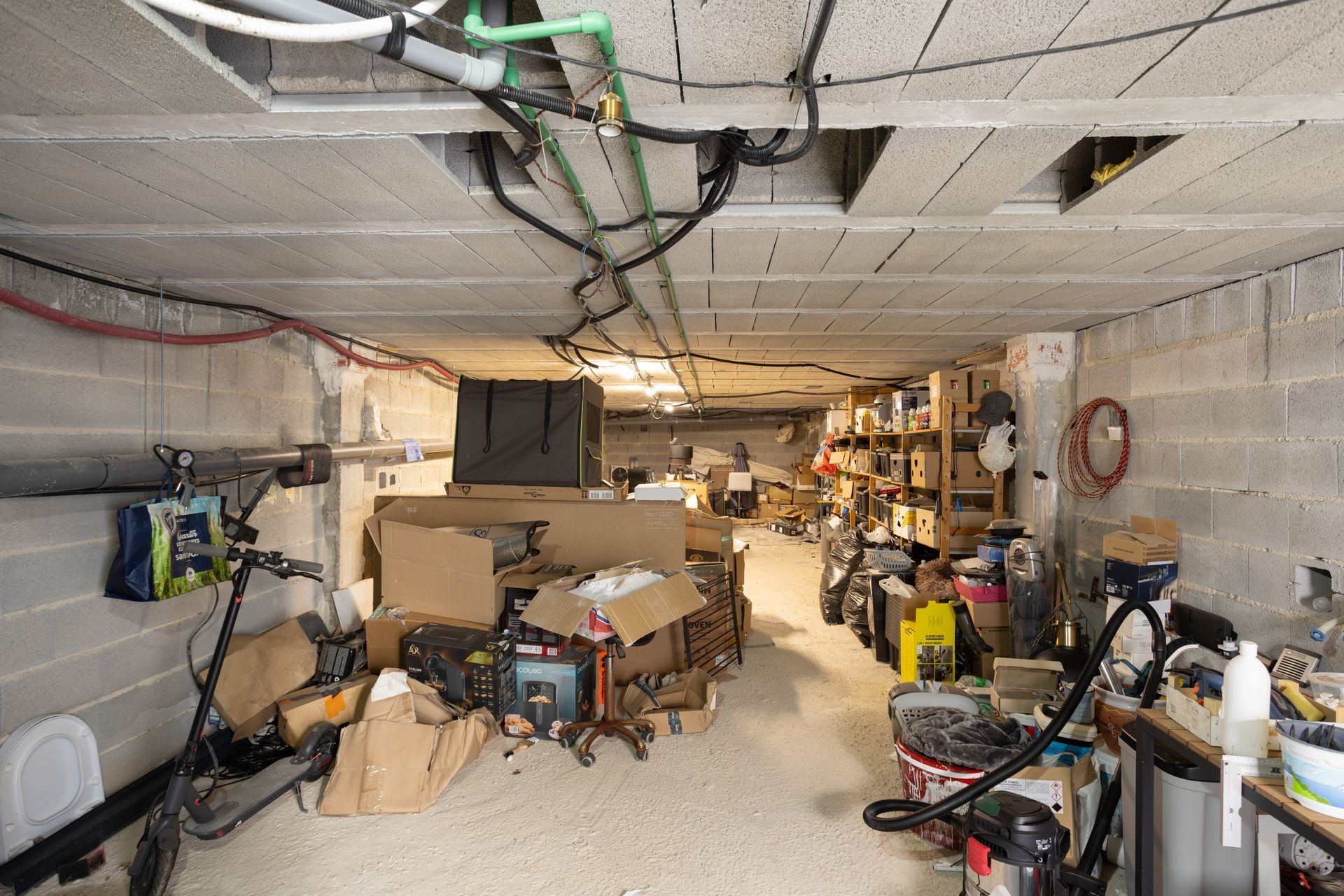 Cluttered basement workshop with boxes, tools, bikes, and exposed ceiling pipes