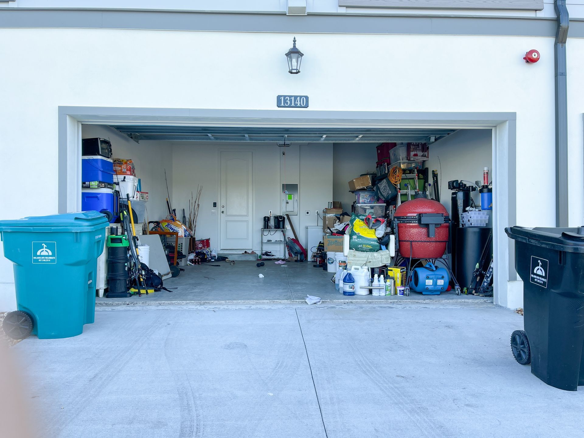 Open garage packed with storage bins, tools, and household items, with trash and recycle bins outside