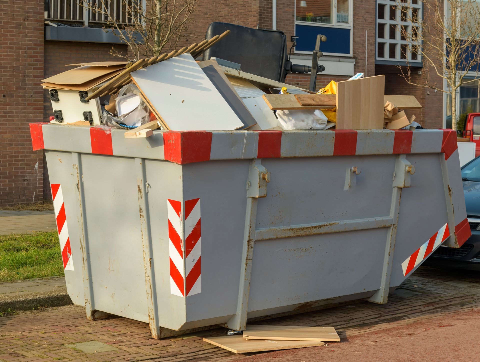 Large gray dumpster filled with construction debris, with red-and-white reflective stripes outdoors.