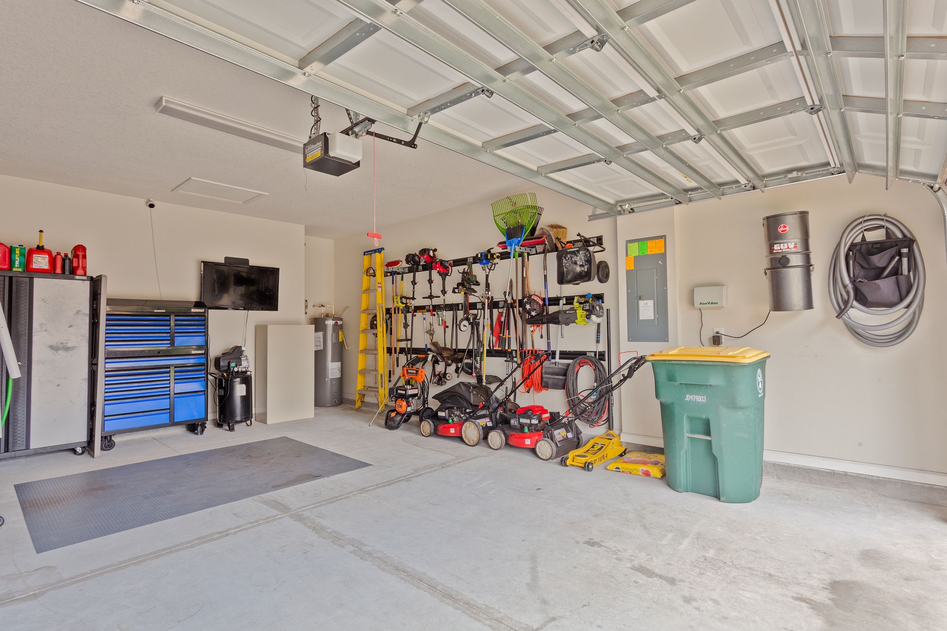 Spacious garage interior with tools, shelving, a ladder, and a green trash bin on a concrete floor