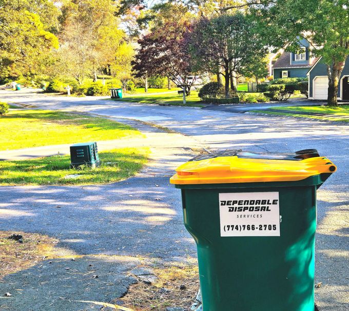Two people place a green trash bag into a large outdoor bin on a sunny day.