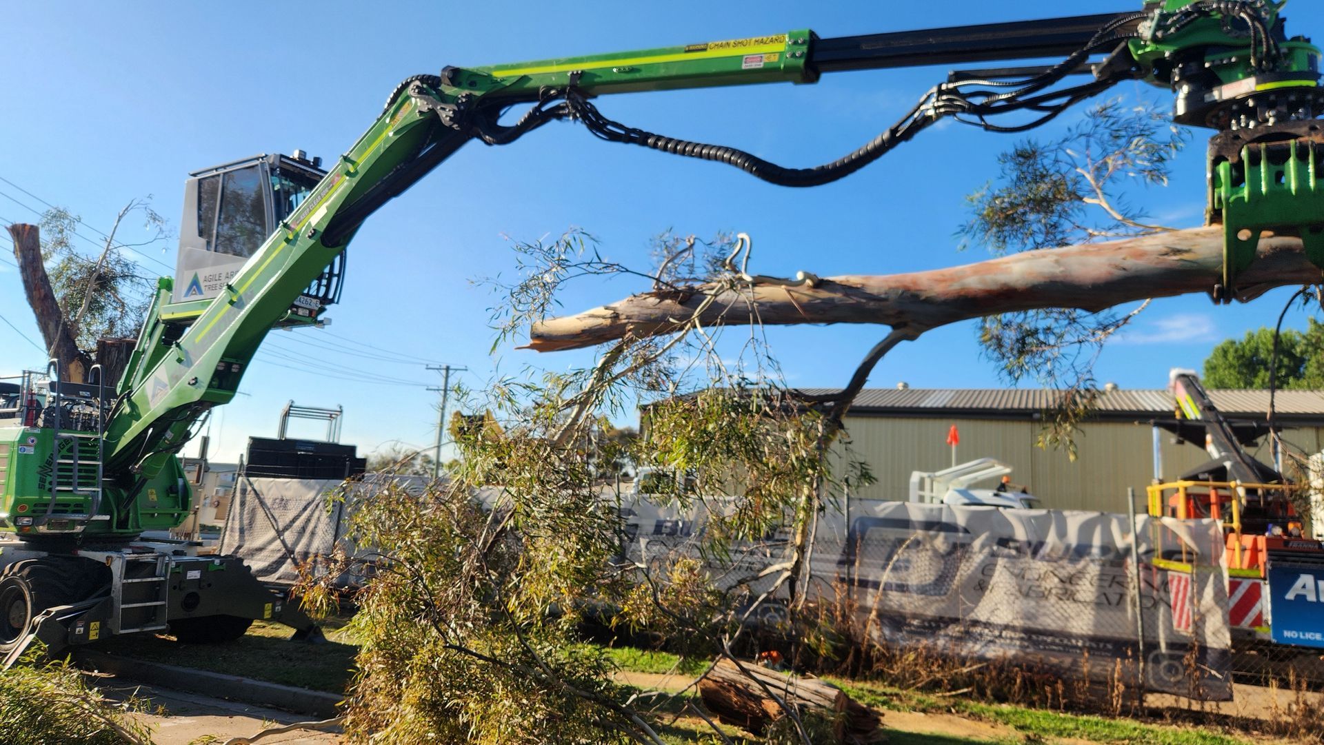 A green forestry crane arm holds a large, horizontal tree trunk in front of a construction site under a clear blue sky.