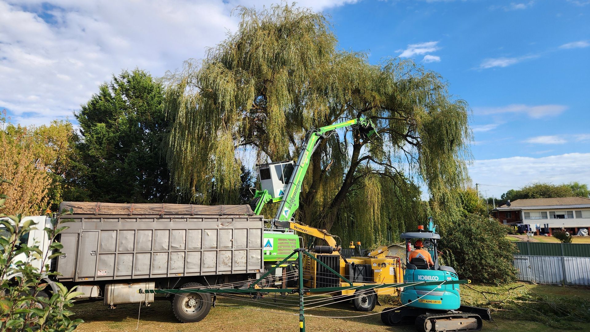 A green mechanical tree shear works on a large willow tree next to a dump truck and a teal mini excavator in a grassy yard.