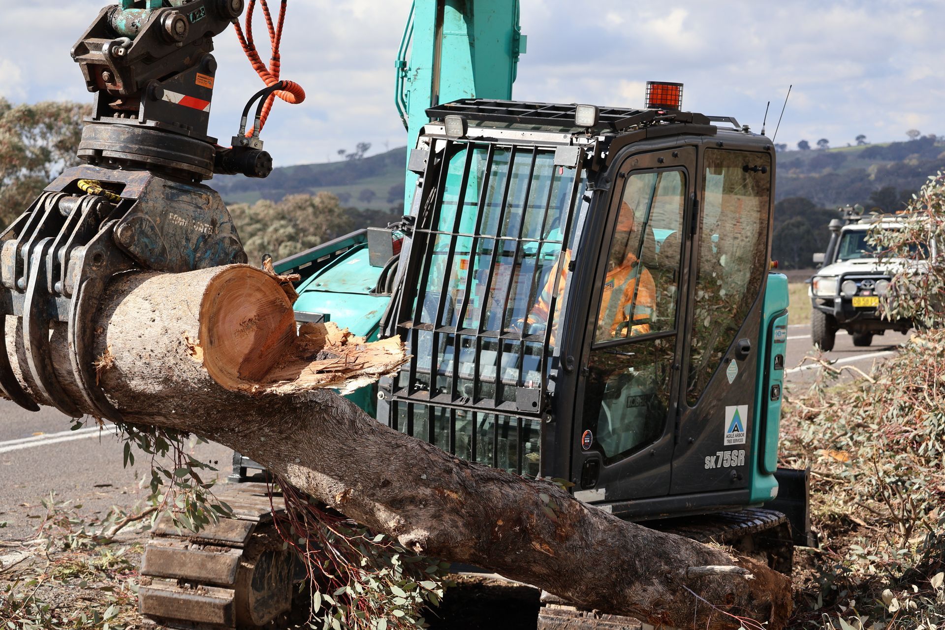 Excavator with claw arm gripping a large tree log, clearing debris alongside a road — Agile Arbor Pty Limited in Kelso, NSW
