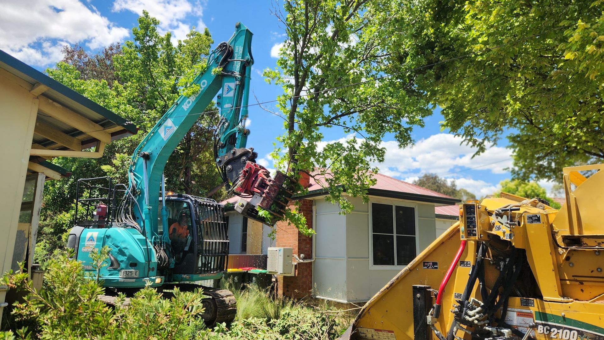 A turquoise excavator and yellow wood chipper work to clear tree branches near a house under a blue, cloudy sky.