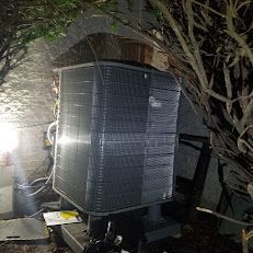 A large air conditioner is sitting under a tree in a shed.