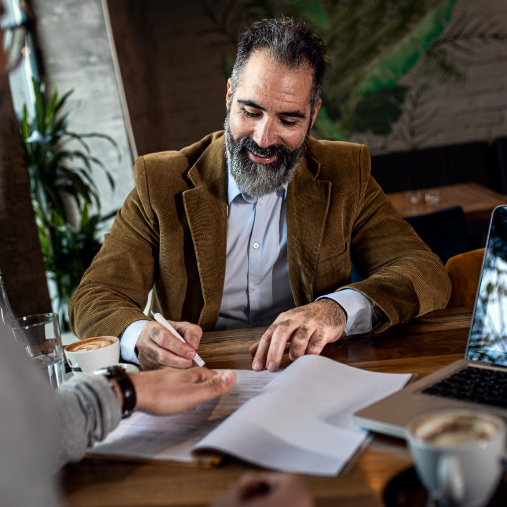A man with a beard is sitting at a table signing a document.