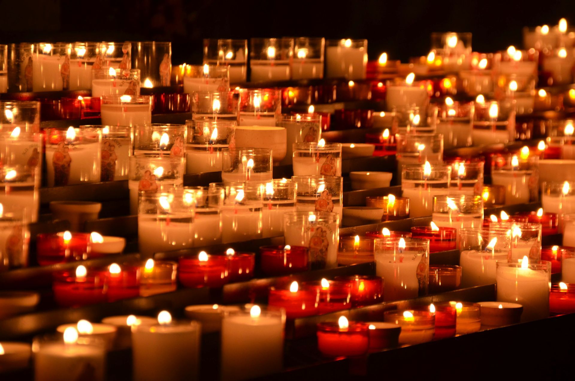 Rows of glowing votive candles in glass holders, illuminating a dark, serene setting.