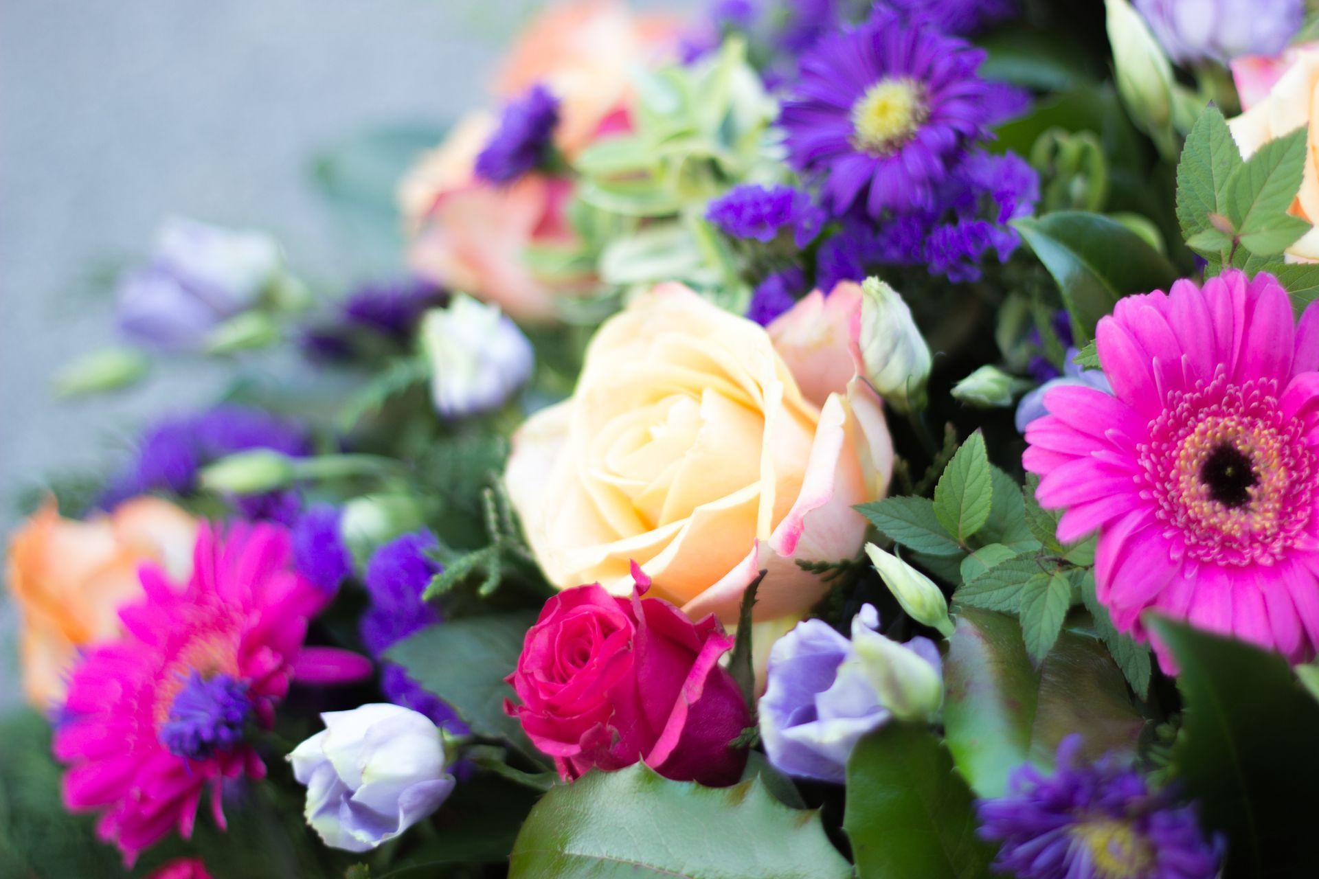 Bouquet of mixed flowers with a pale yellow rose, pink gerbera daisies, purple blooms, and green leaves