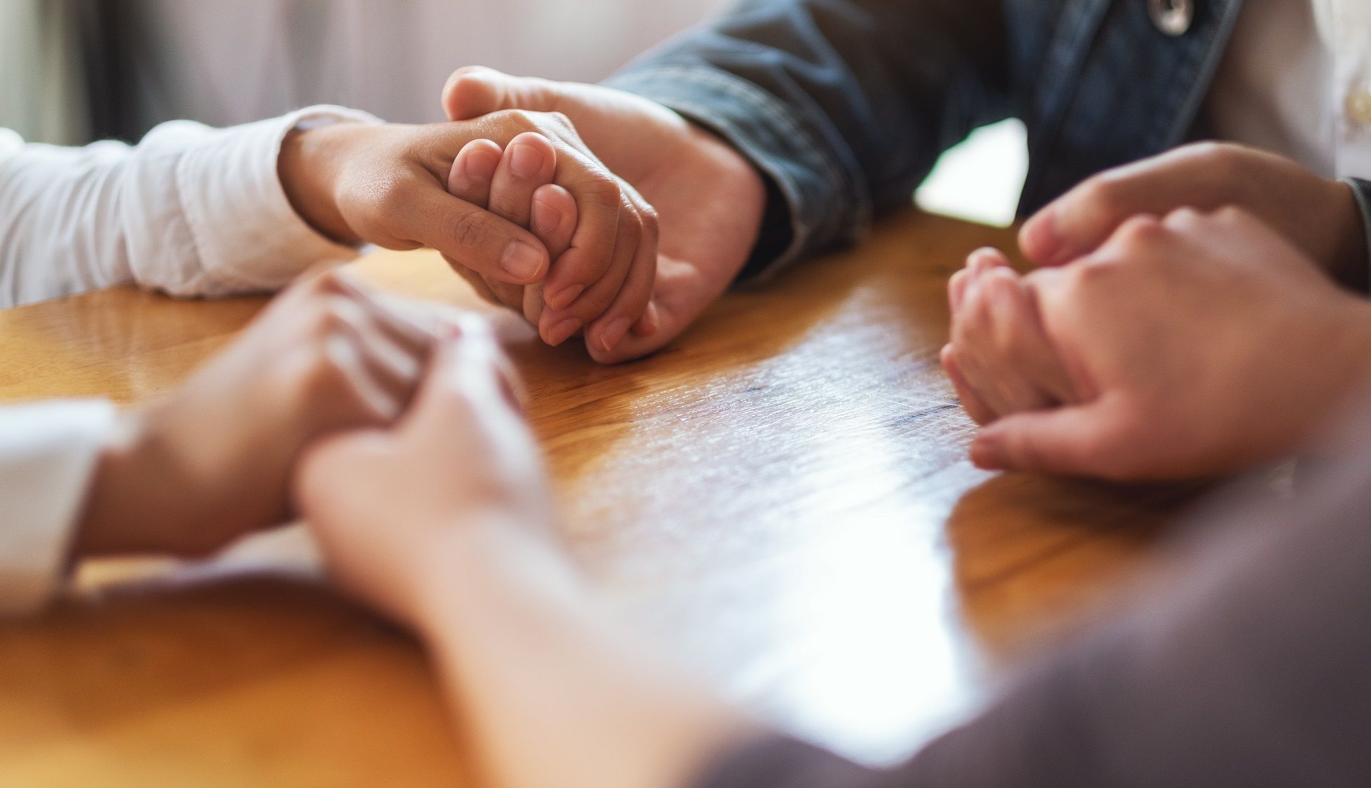 Hands clasped together around a wooden table in a warm indoor group discussion