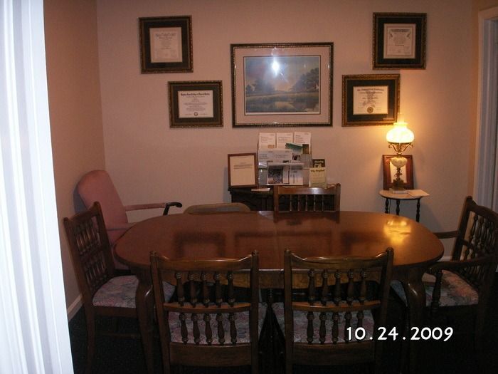 Dining room with oval table, wooden chairs, framed wall art, and a lit lamp on a side table.