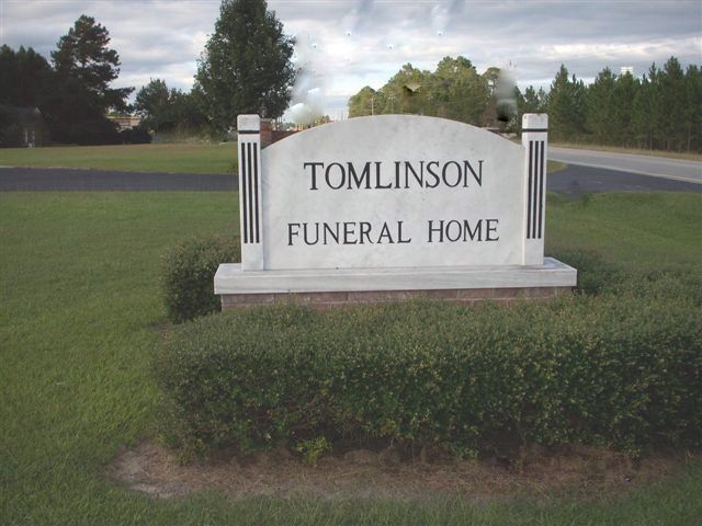 Tomlinson Funeral Home sign on a grassy roadside, with trees and cloudy sky in the background