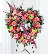 Heart-shaped pink and red floral wreath on a stand with greenery and soft white curtains