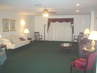 Spacious living room with green carpet, white sofa, lamps, chairs, and curtained window.
