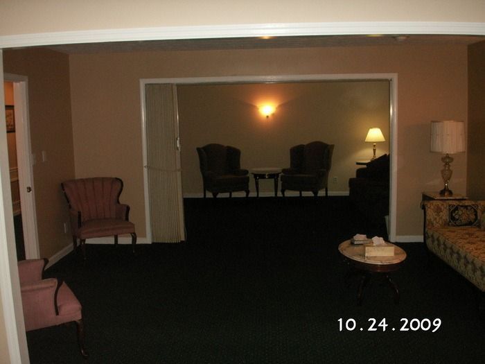 Dim hotel room with chairs, lamp, patterned sofa, and a dark recessed seating area.