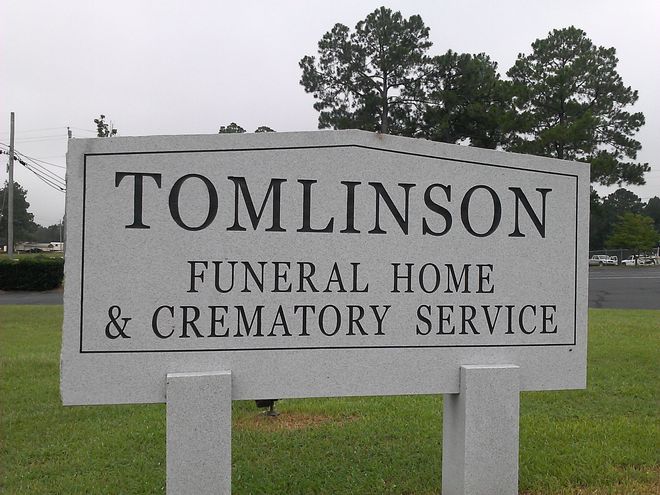Tomlinson Funeral Home & Crematory Service sign on a gray monument sign by a grassy roadside.