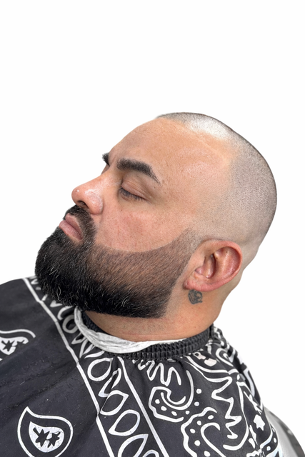 Side profile of a man with a closely shaved head and a neatly trimmed, sharp-edged dark beard wearing a patterned cape.