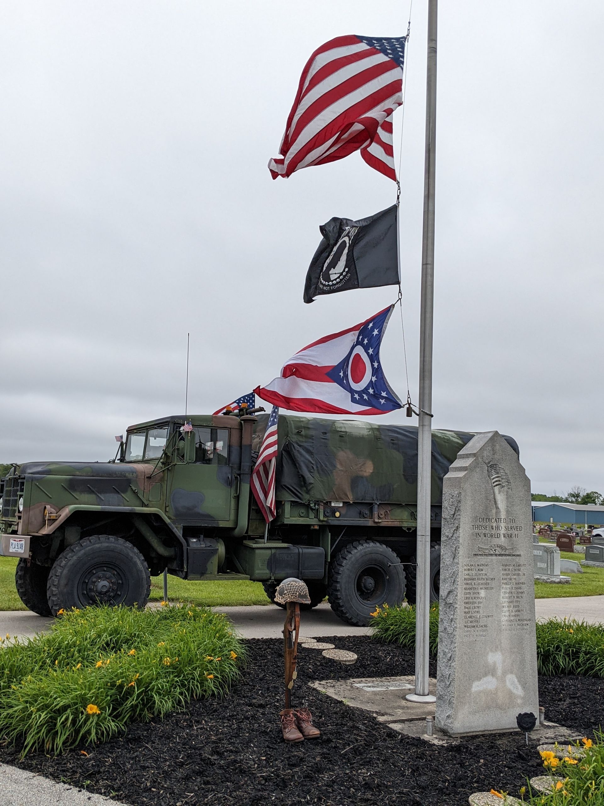 Military truck, flags (US, POW/MIA, Ohio), memorial stone, and foliage. Cloudy sky.